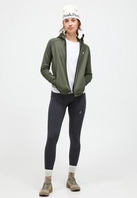 Olive green zip-up jacket over a white t-shirt, paired with black leggings and beige hiking boots. The jacket has a black zipper.