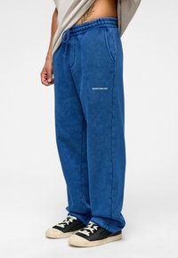 Blauwe trainingsbroek van zacht materiaal met een elastische tailleband en trekkoord. Voorzien van zijzakken en de tekst "MISSIE VOLTOOID." in het wit.