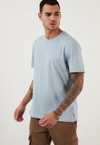 Tricou din bumbac albastru, cu guler rotund și mâneci scurte. Are o croială dreaptă, textură fină și fante laterale. Este purtat cu pantaloni scurți cargo bej.