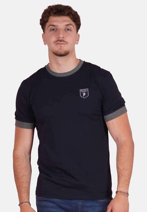 Jeune homme aux cheveux courts et bouclés portant un T-shirt noir avec col et bordures de manches grises, debout devant un fond uni.