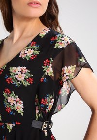 Robe noire à motif floral avec des manches courtes en voile ; présente des motifs floraux colorés en rose, rouge et vert ; taille cintrée avec une boucle de ceinture.