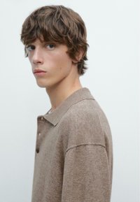 Polo shirt marrone in maglia testurizzata con colletto, caratterizzato da una vestibilità rilassata e una chiusura con un bottone, indossato da un modello con capelli ondulati.