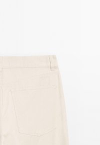 Vue arrière d'un pantalon beige montrant la ceinture, les passants de ceinture et une poche arrière unique avec des coutures visibles sur un fond uni.