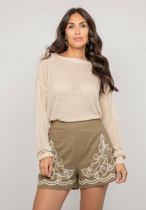 Femme aux longs cheveux bruns portant un pull en maille beige et un short vert olive avec des motifs floraux brodés blancs, debout devant un fond uni.
