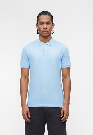 Jeune homme aux cheveux tressés portant un polo bleu clair à manches courtes et un pantalon foncé, se tenant devant un fond blanc uni.