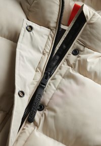 Beige dunjakke med sort lynlås med branding, trykknapper og en orange detalje inde i kraven. Blød, quiltet tekstur.