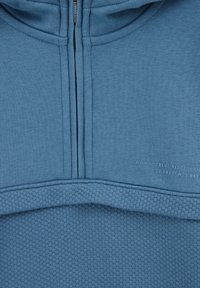 Hoodie azzurro chiaro con sezione inferiore texturizzata, tessuto superiore liscio, zip frontale e marchio stampato sul petto.