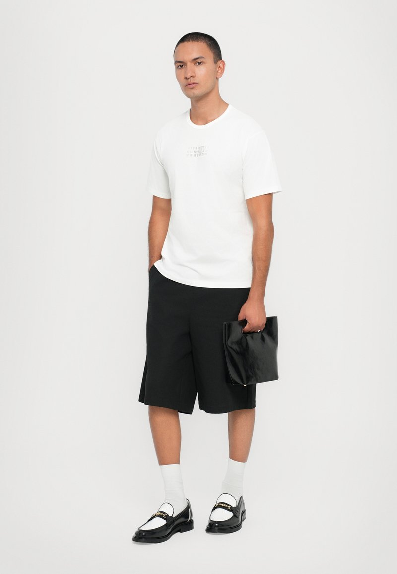 T-shirt bianco in cotone con dettagli grafici, pantaloni corti neri su misura, pochette nera in pelle, calzini bianchi e mocassini neri con accenti bianchi.