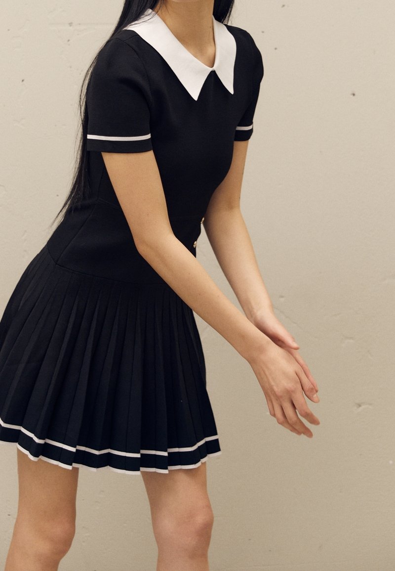 Femme portant une robe plissée noire avec un col blanc et des bordures aux manches blanches, debout les mains jointes devant un fond uni.