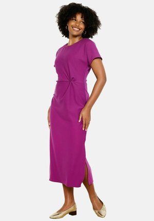 Lokkis juustega naine, kes kannab põlveni ulatuvat lilla kleiti küljelõhikuga ja kuldsete madala kontsaga kingi, naeratab ja vaatab kõrvale.