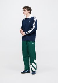 Μπλε πόλο μπλούζα με άσπρο κολάρο και ριγέ μανίκια, συνδυασμένη με πράσινο παντελόνι με άσπρες διαγώνιες λεπτομέρειες. Οι μπλε αθλητικά παπούτσια ολοκληρώνουν την εμφάνιση.