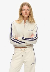 Sweatshirt crème zippé avec des manches rayées marines, affichant un logo sur la poitrine et un pantalon de survêtement crème assorti avec taille à cordon.