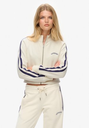 Sudadera cremosa con cremallera y mangas rayadas en azul marino, con un logo en el pecho y pantalones de chándal cremosos a juego con cintura con cordón.