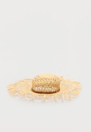 Chapeau en paille tressée à larges bords avec bords frangés et motif ajouré autour de la calotte, présenté sur un fond uni.