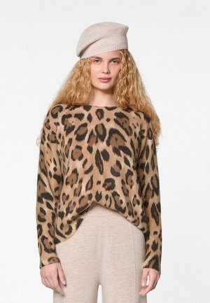 Vrouw met lang krullend haar, gekleed in een beige baret, een trui met luipaardprint en beige broek, staand tegen een effen witte achtergrond.