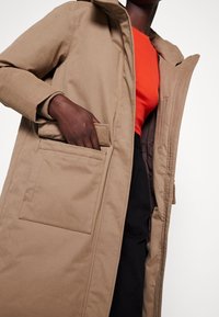 Beige yderjakke med hætte, stor frontlomme og ribbede manchetter. Bæres over en lys orange top med en glat tekstur.