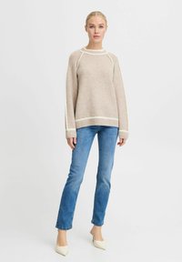 Beige gebreide trui met witte strepen bij de kraag en zoom, gecombineerd met blauwe spijkerbroek en lichtgekleurde schoenen, met een relaxte pasvorm.