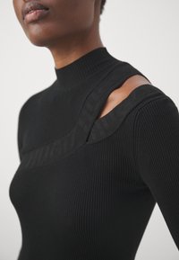 Pull noir côtelé avec col haut et découpe aux épaules. Comprend une bande de logo texturée traversant la couture de l'épaule.