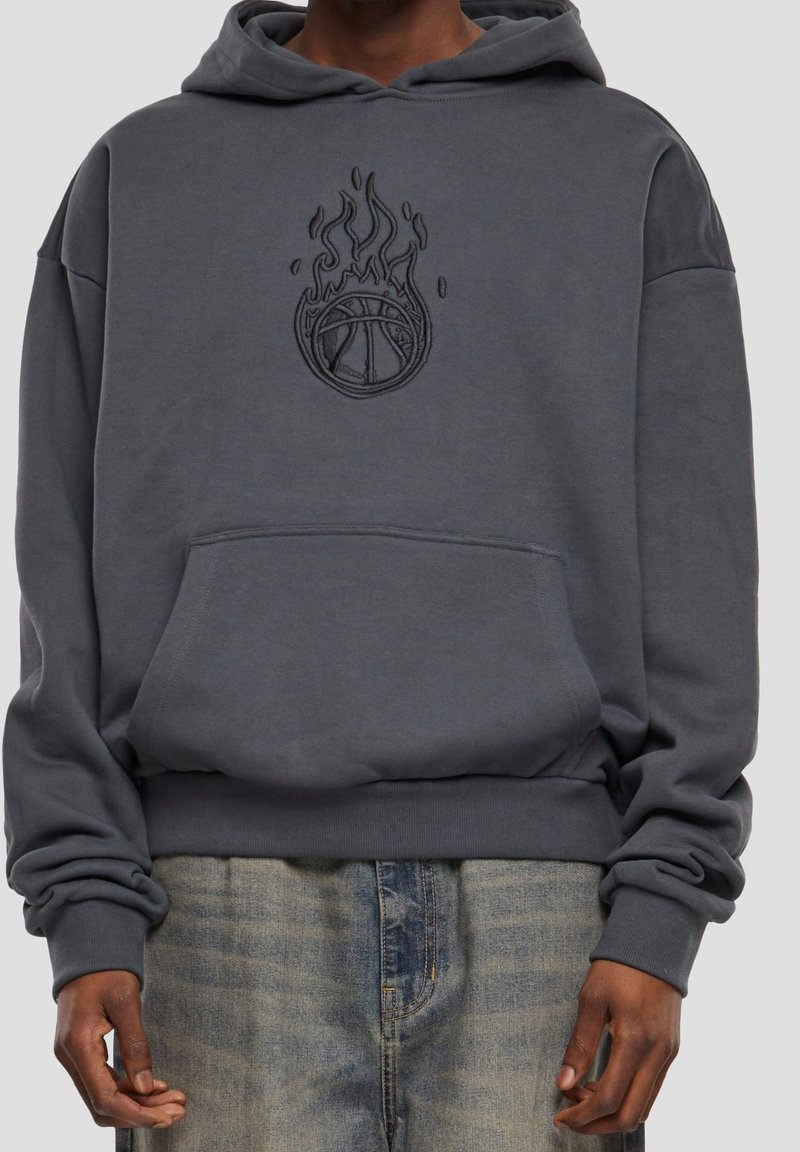 Personne portant un sweat à capuche gris foncé avec une poche avant et un graphisme noir de ballon de basket enflammé sur la poitrine, associé à un jean bleu délavé.