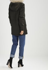 Tmavě zelená parka s umělou kožešinou na kapuci, s zipem a patentky, nošena přes zmačkané modré džíny a černé lodičky.