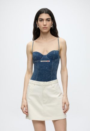 Frau mit langen dunklen Haaren trägt ein blaues Denim-Korsett-Top und einen beigen Rock, steht vor einem schlichten hellen Hintergrund.