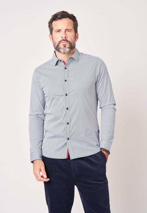 Man met baard die een lichtgekleurde, langermelige blouse met patroon en donkere broek draagt, staand met één hand in zijn zak tegen een effen achtergrond.