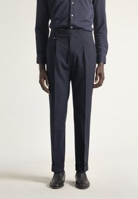 Pantalones ajustados de color marino con una textura suave, que presentan una cinturilla alta, pliegues laterales y acentos de botón. Combinados con zapatos negros.