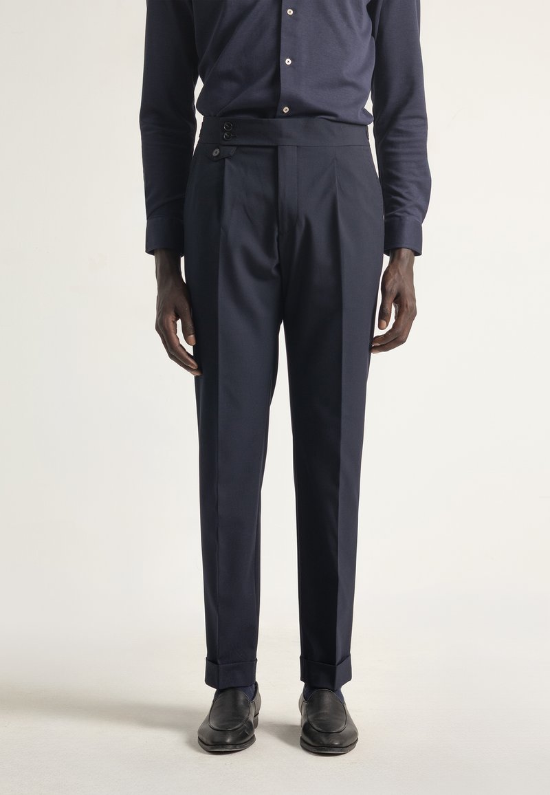 Pantalones ajustados de color marino con una textura suave, que presentan una cinturilla alta, pliegues laterales y acentos de botón. Combinados con zapatos negros.