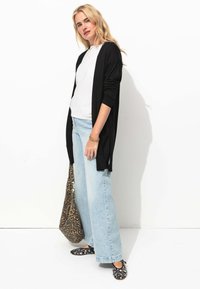 Un long cardigan noir sur un haut blanc, associé à un jean clair à jambes larges et des chaussures noires à fleurs. Elle tient un sac à imprimé léopard.