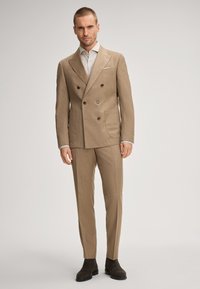 Dunkelbrauner Anzug aus strukturiertem Stoff mit doppelter Knopfleiste, zwei großen Fronttaschen, braunen Knöpfen und passenden, schmal geschnittenen Hosen, kombiniert mit dunklen Stiefeln.