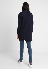 Tmavě modrý dlouhý kardigan s texturovaným vzorem pletení, volným střihem a postranními rozparky, kombinovaný s úzkými modrými džíny a bílými teniskami.