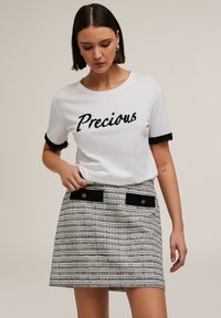 Camiseta blanca con la palabra "Precious" en letras texturizadas negras, combinada con una falda a rayas con un patrón que presenta acentos negros y detalles en dorado.