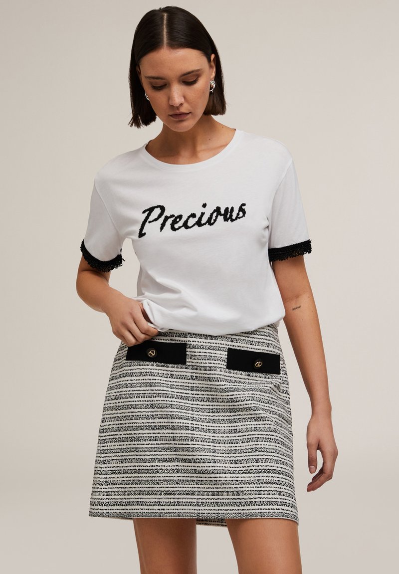 Camiseta blanca con la palabra "Precious" en letras texturizadas negras, combinada con una falda a rayas con un patrón que presenta acentos negros y detalles en dorado.