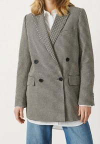 Blazer croisé en tissu à motif pied-de-poule noir et blanc. Il comporte deux poches avant et des boutons noirs.
