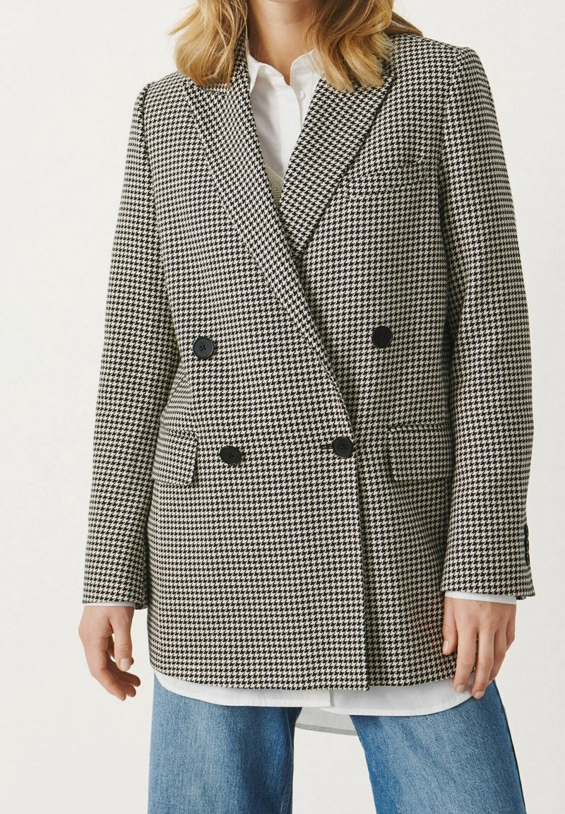 Blazer croisé en tissu à motif pied-de-poule noir et blanc. Il comporte deux poches avant et des boutons noirs.
