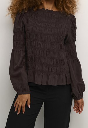 Žena v dlouhém rukávu tmavě hnědé nabírané blůze s elastickými manžetami a černých kalhotách, stojící před jednoduchým pozadím.