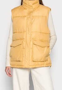 Gelbe, gepolsterte Weste mit hohem Kragen, vorderen Druckknöpfen und zwei großen aufgesetzten Taschen. Glatte Textur mit matter Oberfläche.