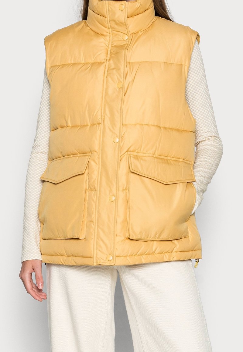 Gelbe, gepolsterte Weste mit hohem Kragen, vorderen Druckknöpfen und zwei großen aufgesetzten Taschen. Glatte Textur mit matter Oberfläche.