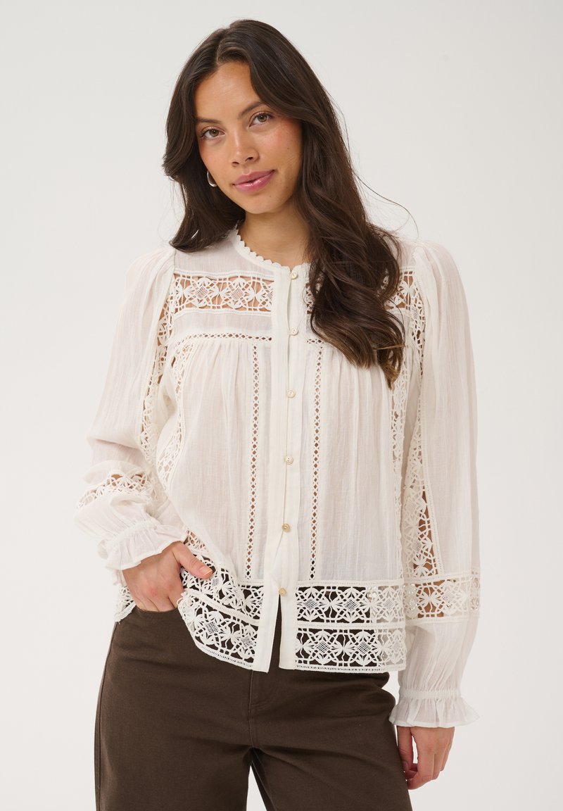 Femme aux longs cheveux bruns, portant un chemisier blanc orné de dentelle et un pantalon marron, debout devant un fond clair uni.
