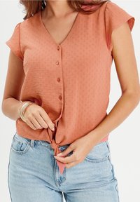 Haut à manches courtes de couleur pêche avec col en V, détails de boutons, tissu texturé et nœud noué à la taille, assorti à un jean bleu clair.