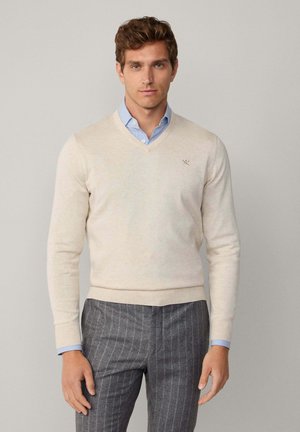 V-hals beige sweater lavet af blødt materiale med ribbede manchetter og kant, båret over en lyseblå stribet skjorte.