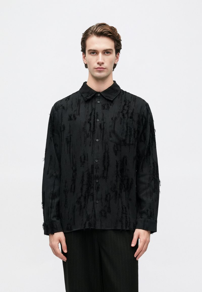 Νέος άνδρας φορώντας ένα μαύρο πουκάμισο με υφή και πέτο, στέκεται μπροστά σε ένα απλό λευκό φόντο.