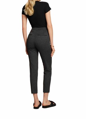 Kvinna med långt blont hår iförd en svart kortärmad topp, figurnära grå byxor som slutar vid anklarna, och svarta slip-on sandaler, sedd bakifrån.