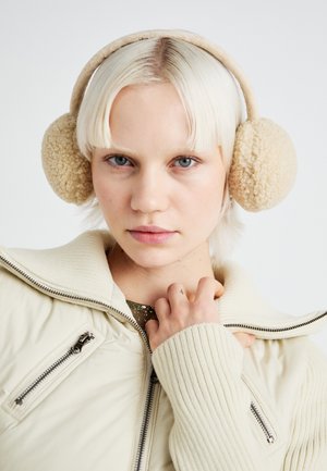 Beige øreklokker laget av fluffy materiale, rund form og myk tekstur. Brukt med en lysfarget, glidelås, stripete jakke med høy krage.