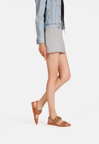 Sandali in pelle marrone con elementi in metallo decorativi, dotati di una suola bianca spessa. Indossati con una gonna grigia chiara e una giacca di jeans.