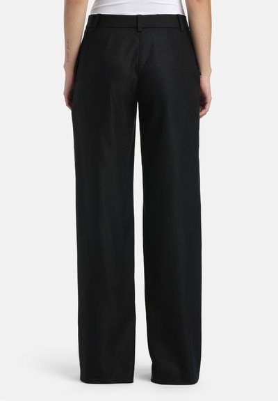 Personne portant un pantalon noir à jambes larges et un haut blanc, debout, vue de dos sur un fond uni.