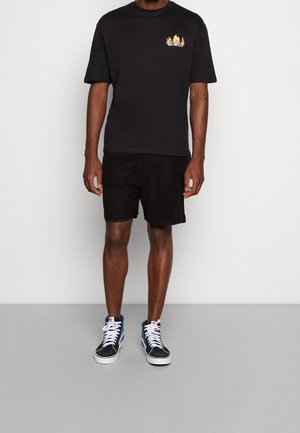Schwarzes T-Shirt mit kurzen Ärmeln und Flammenmotiv auf der linken Brust, kombiniert mit schwarzen Mesh-Shorts und blauen High-Top-Sneakern mit weißen Schnürsenkeln.
