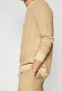 Beige síťovaná košile s dlouhým rukávem a žebrovanými manžetami a lemem. Tkanina je prodyšná, s volným střihem a minimálním značením.