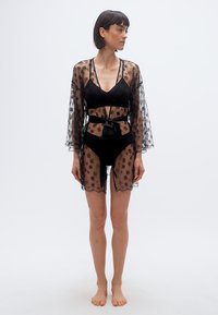 Γυναίκα που φορά διάφανο μαύρο ρόμπα με λουλουδένια δαντέλα δεμένη στη μέση πάνω από μαύρα εσώρουχα, όρθια ξυπόλητη μπροστά σε απλό φωτεινό φόντο.