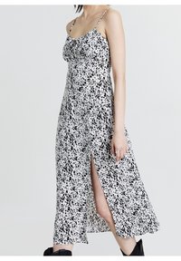 Robe à pois noirs et blancs avec fines bretelles, encolure à volants et fente latérale. Le tissu est léger avec une texture lisse.